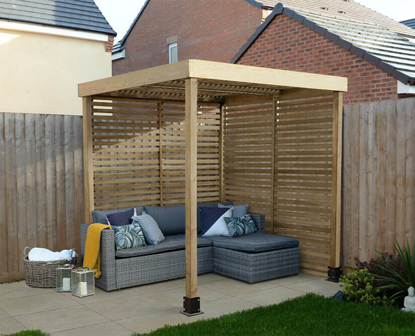 A wooden pergola with slatted panels on 2 adjoining sides, situated in the corner of a garden and furnished with a sofa and cushions