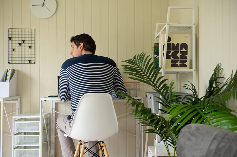 a man works at his desk in a garden office. There is a bookshelf to his right and a plant behind him. A clock on the wall in front of him shows the time 12:45
