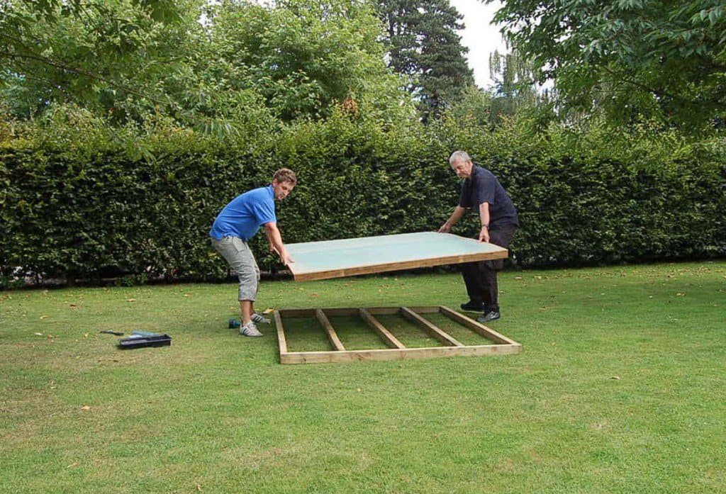 Two men laying a shed base on a grassy area