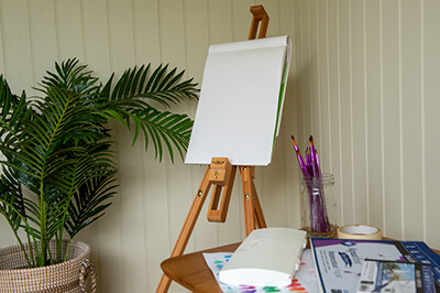an easel set up in the corner of a garden building. a plant to the left of the image. in the immediate foreground, a table with drawing implements