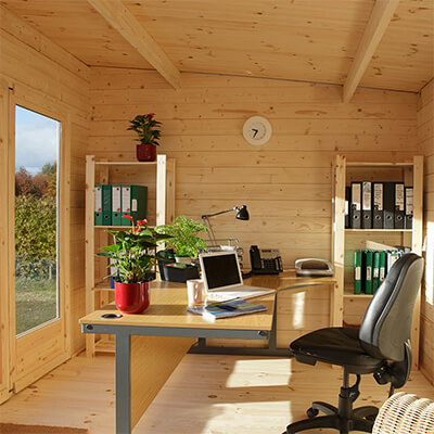 an office setup in a pent log cabin: Desk, laptop chair plant, lamp. Against the far wall are 2 shelving units filled with files