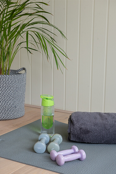 A Possible Use for your Garden Office: A Home Gym: A grey Gym Mat is layed on the floor. On top of it: a green water bottle, a dark grey towel, 2 Grey 1KG Dumbells and 2 smaller Pink Dumbells. There is a potted plant in the background.