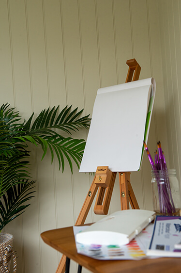 A Possible Use for your Garden Office: An Art Studio: An easel is in the centre of the image with a sketchbook set up on it. In the foreground is a table with brushes and paints on it. In the background is a potted plant.