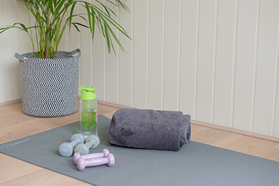 a yoga mat on the floor of a garden building. on top of the mat: a black gym bag, a clear waterbottle with green lid and 4 small dumbells. In the corner of the room is a potted plant