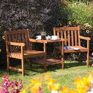 The Rowlinson Hampton Wooden Garden Companion Seat, situated in a beautiful garden, with a magazine on one of the chairs and a tea pot and tea cups on the table.