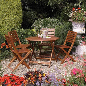 The Rowlinson Plumley 4 Seater Wooden Garden Table and Chairs, situated on gravel, in a beautiful garden, with an ice bucket containing bottles on top of the table.